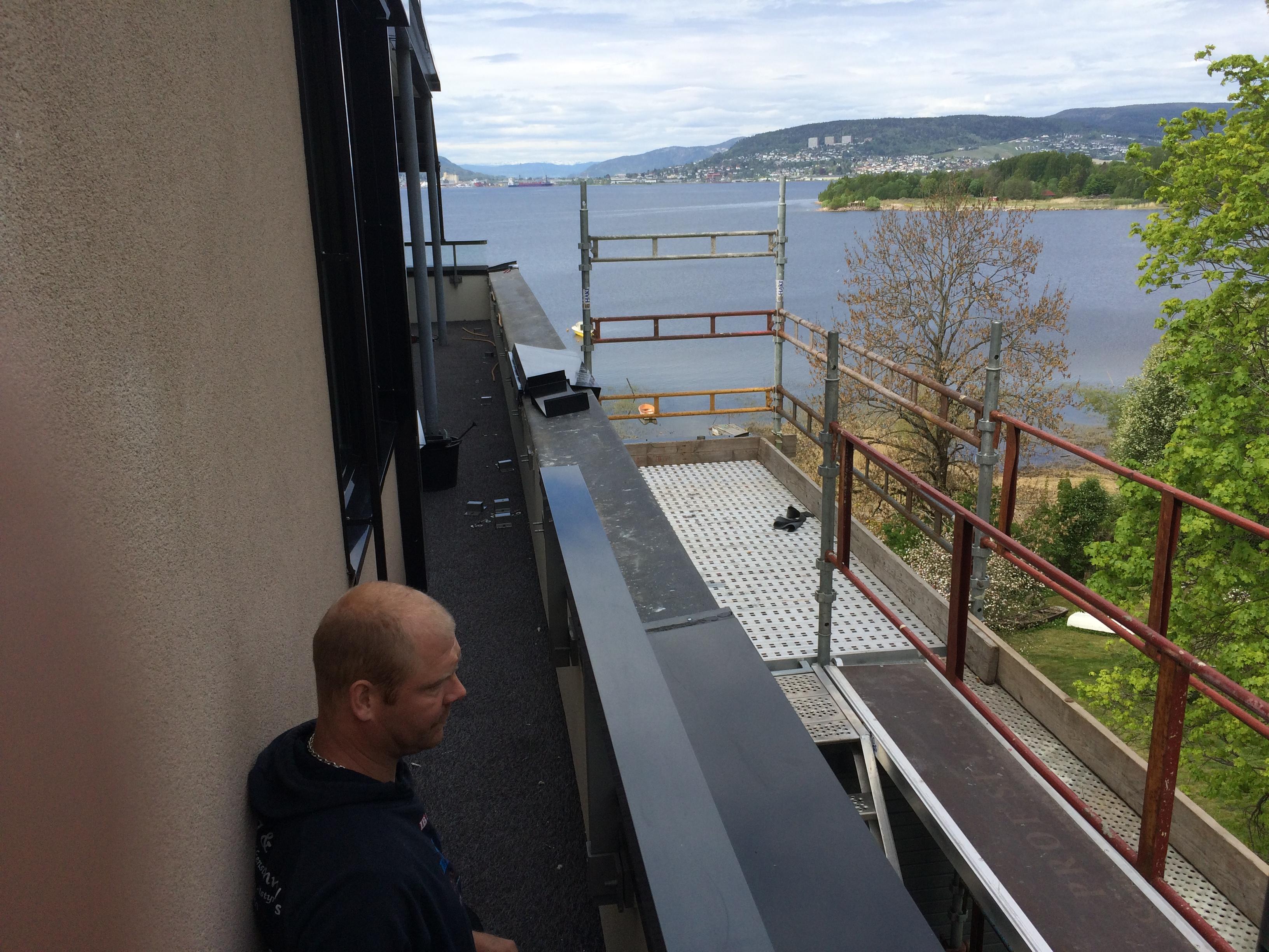 Blikkenslager på stillas ved en ny fasade, med fjorden og åsene bak